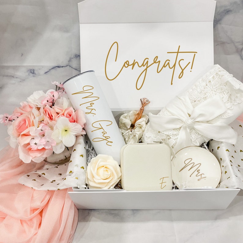 May include: A white gift box filled with a variety of items, including a white tumbler with the text "Mrs. Gage" in gold lettering, a white jewelry box with the letter "E" in gold lettering, a white dish with the word "Mrs." in gold lettering, a white satin robe, and a bouquet of pink and white flowers. The box is decorated with gold hearts and the word "Congrats!" in gold lettering.