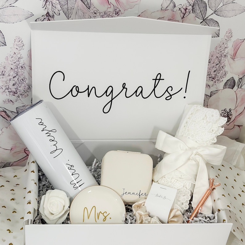 May include: A white gift box with the word "Congrats!" on the lid. The box is filled with various gifts, including a white tumbler with the words "Mrs. [Name]" on it, a white jewelry box with the name "Jennifer" on it, a white satin robe with lace trim, a gold-colored hair scrunchie, and a gold-colored hair clip.