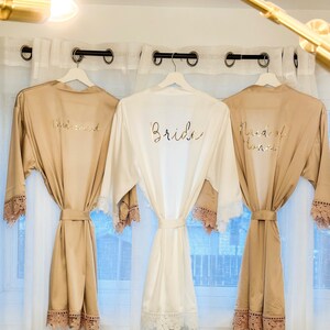 May include: Three satin robes with lace trim, hanging on hangers. The robes are in beige and white, with gold lettering that reads "Bride", "Maid of Honor", and "Bridesmaid".