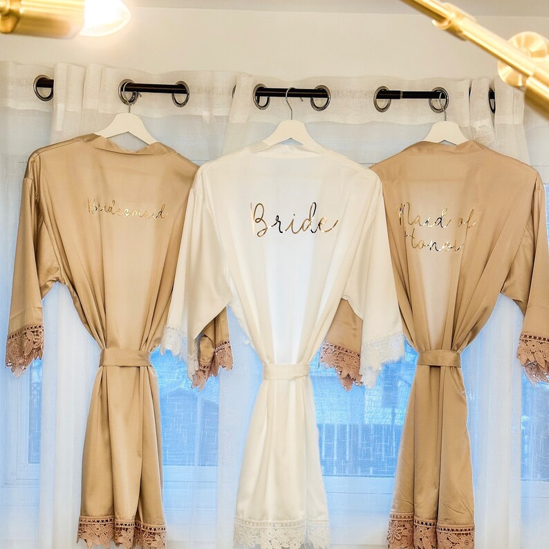 May include: Three satin robes with lace trim, hanging on hangers. The robes are in beige and white, with gold lettering that reads "Bride", "Maid of Honor", and "Bridesmaid".