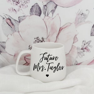May include: White ceramic mug with a curved handle. The mug has the text "Future Mrs. Taylor" in black script, with a small black heart below. The background features a floral pattern with pink and white flowers.