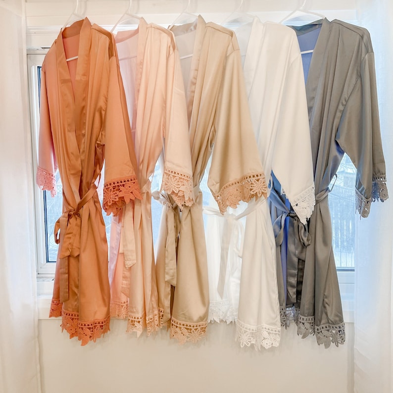 Op de afbeelding: Zes satijnen badjassen met kantversiering in verschillende kleuren, waaronder perzik, beige, wit en grijs. De badjassen hangen aan hangers en hebben een ceintuur.
