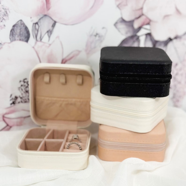 May include: A set of three stackable jewelry boxes. The top box is black with a zipper closure. The middle box is white and the bottom box is beige. All three boxes are square shaped.
