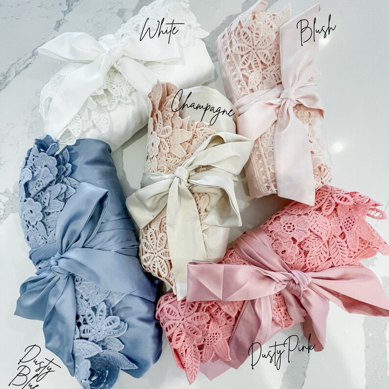 Op de afbeelding: Zes satijnen badjassen met kantversiering in verschillende kleuren, waaronder wit, blush, champagne, stofgrijs en poederroze. Elke badjas is gebonden met een bijpassend satijnen lint.