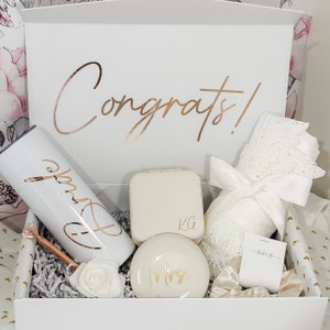 May include: A white gift box filled with bridal shower gifts. The box is decorated with the word "Congrats!" in gold foil. The gifts include a white tumbler with the word "Bride" in gold foil, a white jewelry box with the initials "KG" in gold foil, a white round box with the word "Mrs." in gold foil, a white robe with lace trim, and a white satin ribbon.