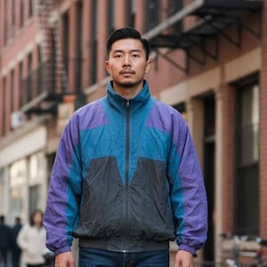 May include: A person wearing a retro nylon jacket in blue, purple, and grey, with dark blue jeans and white trainers. The jacket has a high collar and a full-length zip. The person is walking on a city street, with buildings in the background.