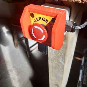 Puede incluir: Un botón rojo de parada de emergencia con fondo amarillo y la palabra "EMERGENCY" impresa en él. El botón está montado en una superficie de madera.