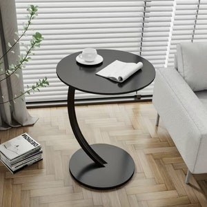 Op de afbeelding: Een zwarte bijzettafel met een gebogen, modern ontwerp. De tafel heeft een rond blad en een ronde basis. De tafel staat op een houten vloer.