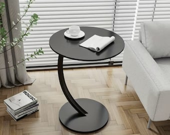 Puede incluir: Una mesa auxiliar negra con un diseño curvo y moderno. La mesa tiene una tapa redonda y una base circular. La mesa está sobre un suelo de madera.