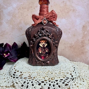 May include: A decorative brown bottle with a textured surface, decorated with a red bow and a jewel. The bottle features an oval frame with a small portrait of a dog. The bottle is placed on a cream-coloured, crocheted doily.