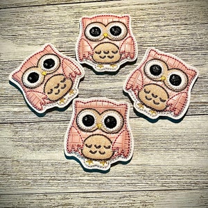 Könnte beinhalten: Vier rosa, eulenformige Aufnäher mit weißen Rändern sind auf einer hellen Holzmaserung angeordnet. Jeder Aufnäher hat schwarze Augen, einen beigen Schnabel und einen strukturierten Körper mit gestickten Details.