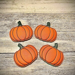 Puede incluir: Cuatro calabazas de fieltro de color naranja con tallos verdes. Las calabazas están dispuestas en forma de cuadrado sobre una superficie de madera marrón claro.