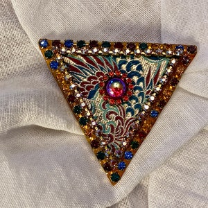 May include: A triangular brooch with a gold-toned frame, adorned with colorful rhinestones and a central iridescent gem. The brooch features a patterned fabric insert with floral motifs in teal, red, and gold. A vintage-inspired accessory.