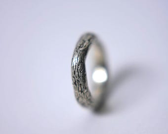 May include: A silver ring with a textured bark-like pattern. The ring is in focus against a blurred white background. The ring's surface has a detailed, organic design, giving it a unique, natural appearance. The ring is a piece of jewelry.