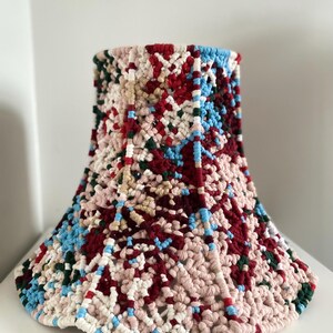 May include: A handcrafted lampshade with a distinctive, textured pattern. The lampshade showcases a vibrant mix of red, blue, green, and cream-coloured yarn. The lampshade is cone-shaped and seems to be made from woven or crocheted material.