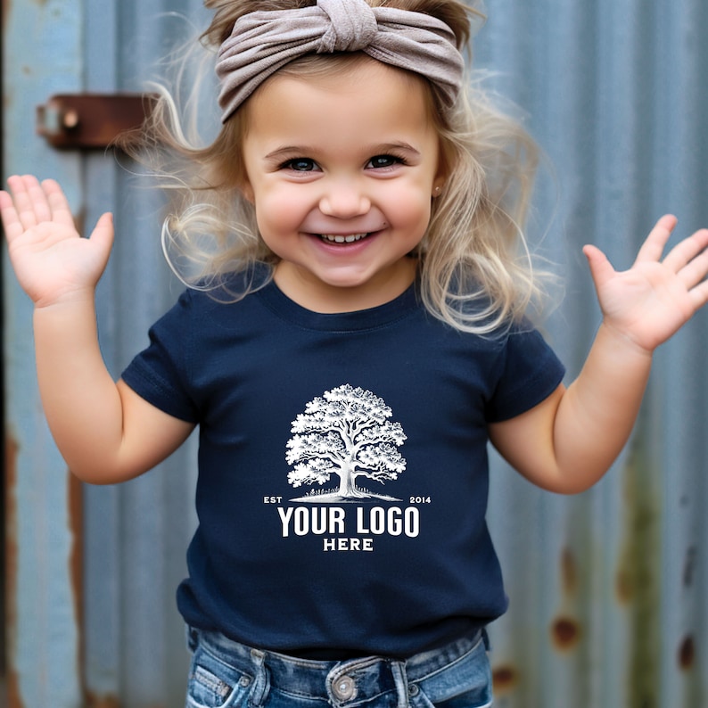 May include: A young child wearing a navy blue t-shirt with a white tree and the text "EST 2014 YOUR LOGO HERE". The child is smiling and has blonde hair with a brown headband.