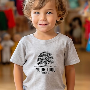 May include: A young child wearing a light gray t-shirt with a black and white tree graphic. The shirt has the text "EST 2014" and "YOUR LOGO HERE" printed on it.