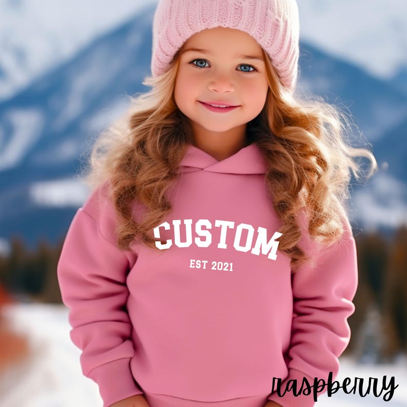 May include: A pink hooded sweatshirt with the text "CUSTOM EST 2021" in white. The sweatshirt is worn by a young girl with blonde hair and a pink beanie.