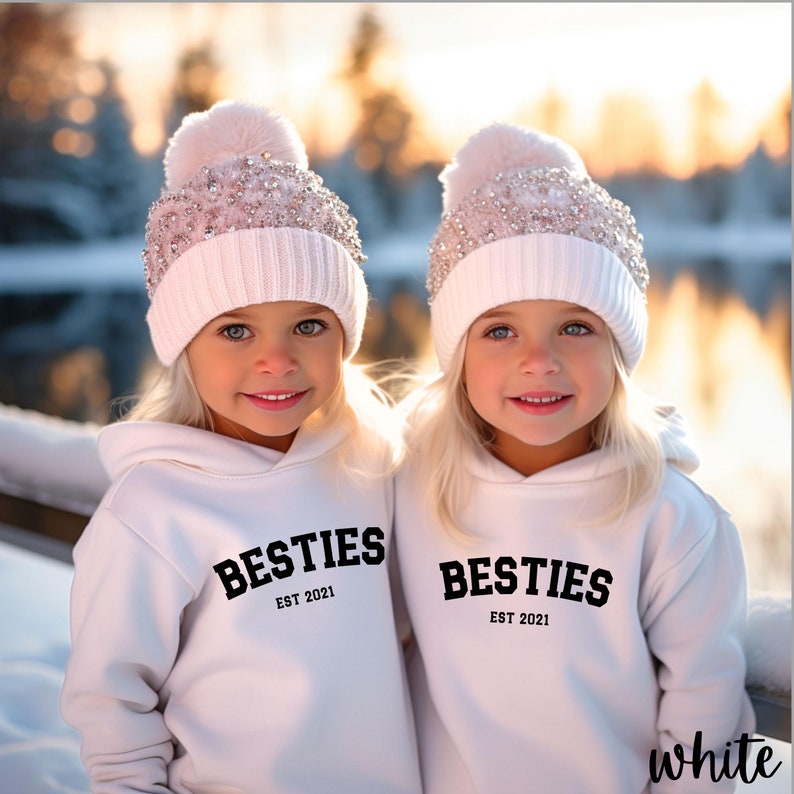 May include: Two children wearing white hoodies with the text "Besties Est 2021" on them. They are both wearing white knit hats with sparkly embellishments and pom-poms.
