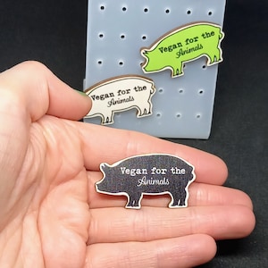 Könnte beinhalten: Drei hölzerne Schweine-Silhouetten mit dem Text "Vegan for the Animals" auf jeder. Die Schweine sind in verschiedenen Farben: grün, weiß und schwarz.