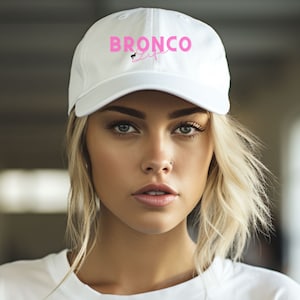 May include: A white baseball cap with a pink embroidered logo that says "BRONCO LIFE". The model is wearing the cap and has blonde hair.