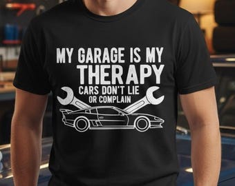 May include: Black t-shirt with the text "MY GARAGE IS MY THERAPY, CARS DON'T LIE OR COMPLAIN" in white. The shirt features a car graphic and wrenches. The person is standing in front of a blue car in a garage.