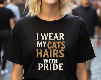 Puede incluir: Camiseta negra con el texto "I Wear My Cats Hairs With Pride" en letras blancas y marrones. La camiseta es de cuello redondo y la lleva una persona con pelo rubio y gafas de sol.