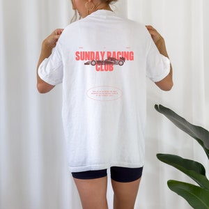 May include: White t-shirt with a red graphic of a race car and the text "SUNDAY RACING CLUB". The text "WHEEL TO WHEEL" is printed above the graphic and the text "JOIN US AS WE WRITE THE NEXT CHAPTER OF RACING HISTORY, ONE LAP AT THE PERFECT LEVEL" is printed below the graphic.