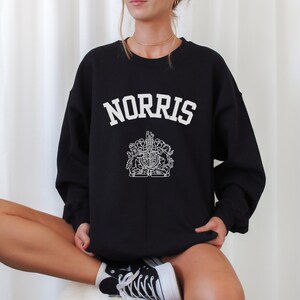 Könnte beinhalten: Schwarzer Pullover mit einem weißen Aufdruck des Wortes "NORRIS" in geschwungener Schrift und einem königlichen Wappen darunter.