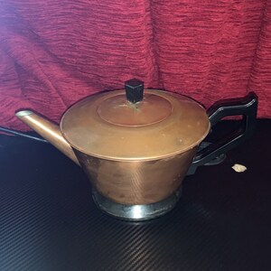 May include: A copper electric kettle with a black handle and lid. The kettle is sitting on a black surface.