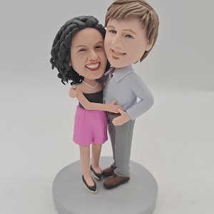 May include: A bobblehead figurine depicting a couple in an embrace. The woman is in a black top and pink skirt, the man in a light blue shirt and grey trousers. They stand on a grey base. The figurine has exaggerated features.