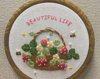 May include: Embroidered hoop art featuring a basket of strawberries, green leaves, and white flowers. The text "BEAUTIFUL LIFE" is stitched above the basket. Two bees and yellow star accents are also included. The hoop is framed in a wooden ring.