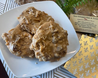 Peut inclure: Plusieurs pralines aux noix de pécan de couleur beige sont disposées sur une assiette carrée blanche. Les pralines ont une surface brillante et texturée avec des morceaux de noix de pécan visibles. L'image montre également une carte avec du texte.
