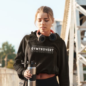 May include: A person wearing a black hooded sweatshirt with the text 'GYMTROVERT' printed on it. The person is holding a silver water bottle and a black gym bag with a red stripe.