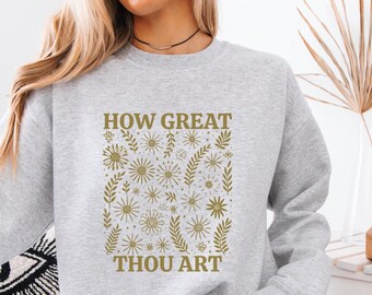 Puede incluir: Una sudadera gris con el texto "HOW GREAT THOU ART" en letras doradas. El texto está rodeado de un patrón de flores y hojas doradas.