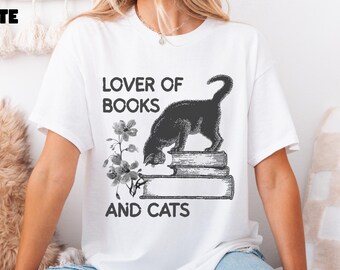 Könnte beinhalten: Weißes T-Shirt mit dem schwarzen Text "LOVER OF BOOKS AND CATS". Eine schwarze Katze ist auf einem Stapel Bücher abgebildet, mit floralen Details. Das Wort "WHITE" befindet sich in der oberen linken Ecke.