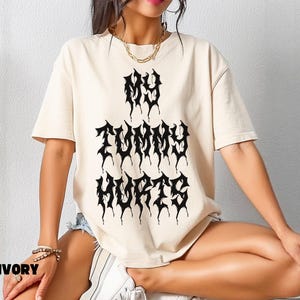 May include: Ivory-colored t-shirt with the text "MY TUMMY HURTS" in a black, gothic font. The shirt has short sleeves and a relaxed fit. The word "IVORY" is printed in a black, bold font at the bottom left corner.