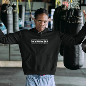 以下が含まれることがあります： 前面に白い文字で「GYMTROVERT」とプリントされた黒いフード付きスウェットシャツ。テキストは縦に積み重ねられており、各単語は上の単語からわずかにオフセットされています。