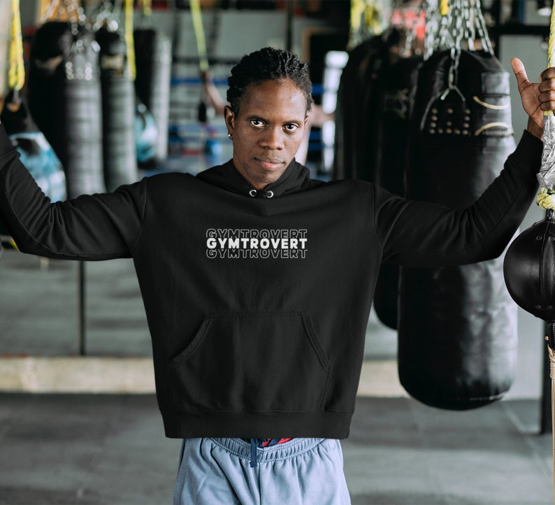 以下が含まれることがあります： 前面に白い文字で「GYMTROVERT」とプリントされた黒いフード付きスウェットシャツ。テキストは縦に積み重ねられており、各単語は上の単語からわずかにオフセットされています。