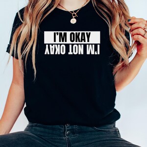 Op de afbeelding: Zwart T-shirt met witte tekst die "Het gaat niet goed met me, het gaat goed met me" zegt.