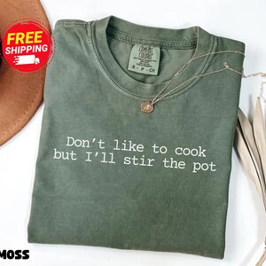 May include: A moss green t-shirt with the slogan "Don't like to cook but I'll stir the pot" in white. The shirt has a crew neck and short sleeves. A gold necklace with a pendant is draped across the shirt.