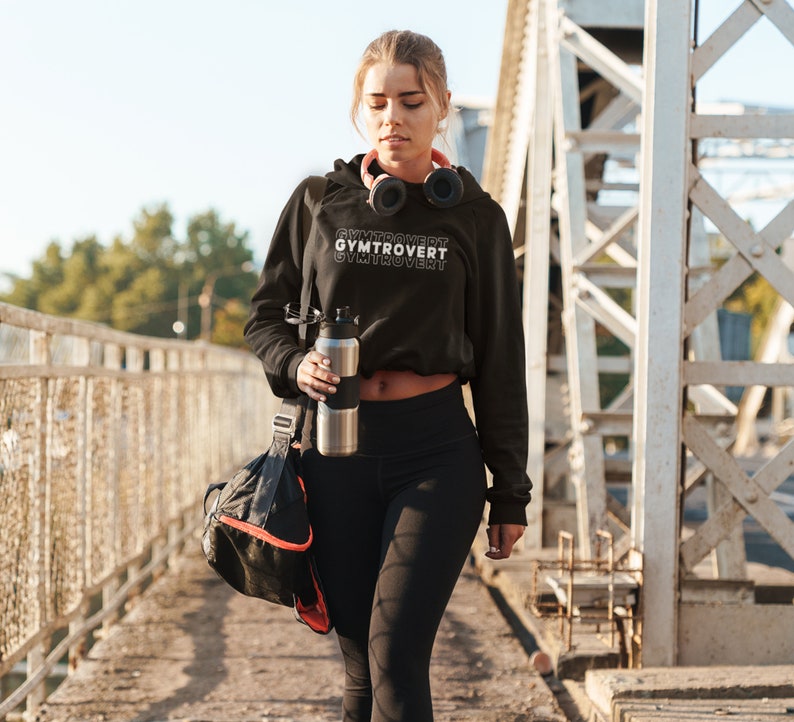 May include: A person wearing a black hooded sweatshirt with the text 'GYMTROVERT' printed on it. The person is holding a silver water bottle and a black gym bag with a red stripe.