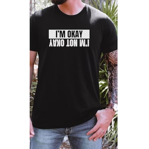 Op de afbeelding: Een zwart T-shirt met witte tekst die "Het gaat niet goed met me. Het gaat goed met me." luidt. De tekst staat in een kader.