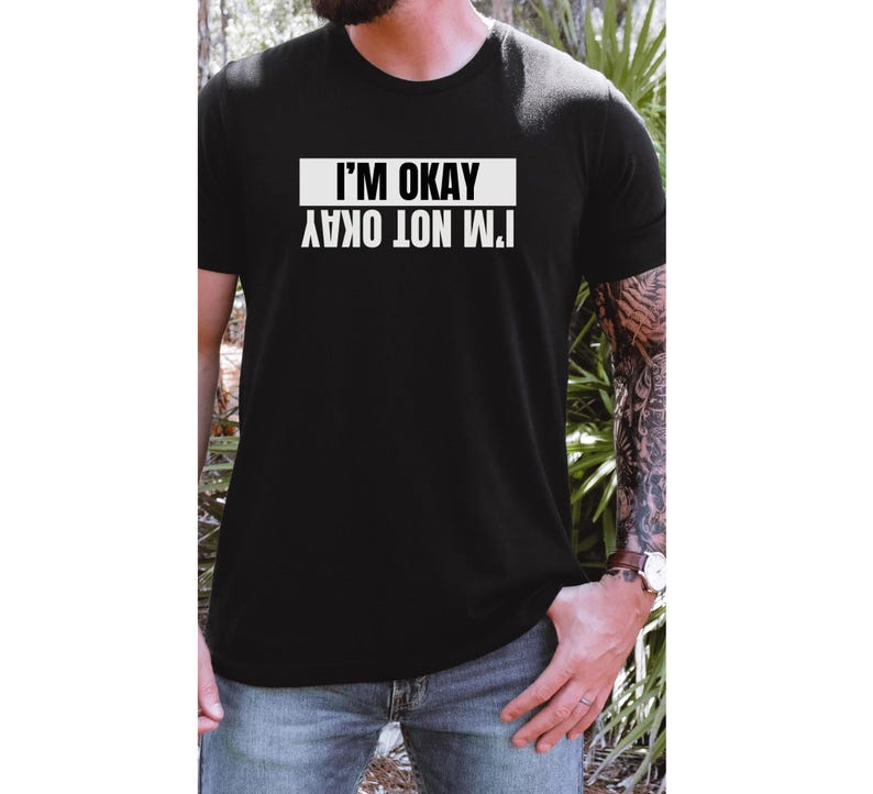 Op de afbeelding: Een zwart T-shirt met witte tekst die "Het gaat niet goed met me. Het gaat goed met me." luidt. De tekst staat in een kader.