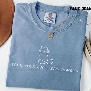 Puede incluir: Una camiseta azul vaquero Comfort Colors con un contorno de gato blanco y el texto "TELL YOUR CAT I SAID PSPSPS". Un collar dorado con un colgante está drapeado sobre la camisa.