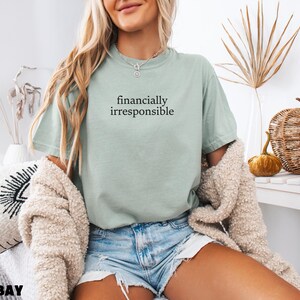 May include: A woman wearing a light green t-shirt with the text "financially irresponsible" printed on it. She is wearing blue denim shorts and a cream-colored cardigan.
