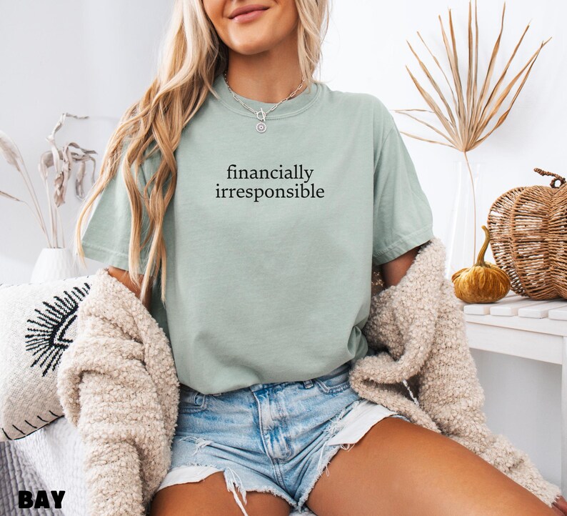 May include: A woman wearing a light green t-shirt with the text "financially irresponsible" printed on it. She is wearing blue denim shorts and a cream-colored cardigan.