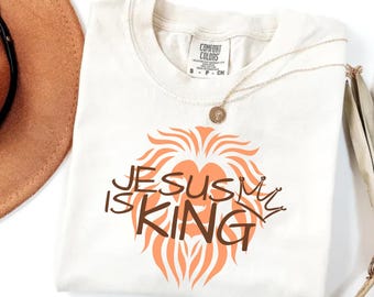 May include: Ivory t-shirt with a lion's mane graphic and the text "Jesus is King" in brown lettering. The shirt has a crew neck and short sleeves.