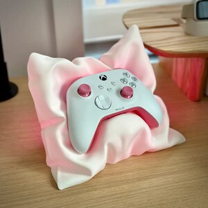 May include: A white Xbox controller with pink accents rests on a pink pillow. The controller has a white faceplate with a black directional pad and a white Xbox logo. The controller is resting on a pink pillow with a white background.