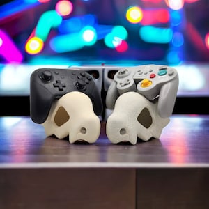Può includere: Due controller per videogiochi, uno nero e uno grigio, poggiano su supporti a forma di teschio. I supporti sono bianco sporco e grigi, con una finitura strutturata. Lo sfondo è sfocato con luci colorate.